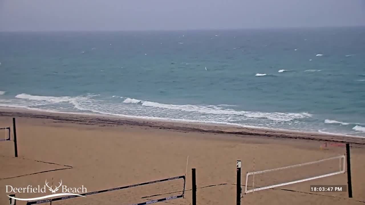 A sandy beach with volleyball nets is shown, overlooking a teal ocean with small waves under an overcast sky.
