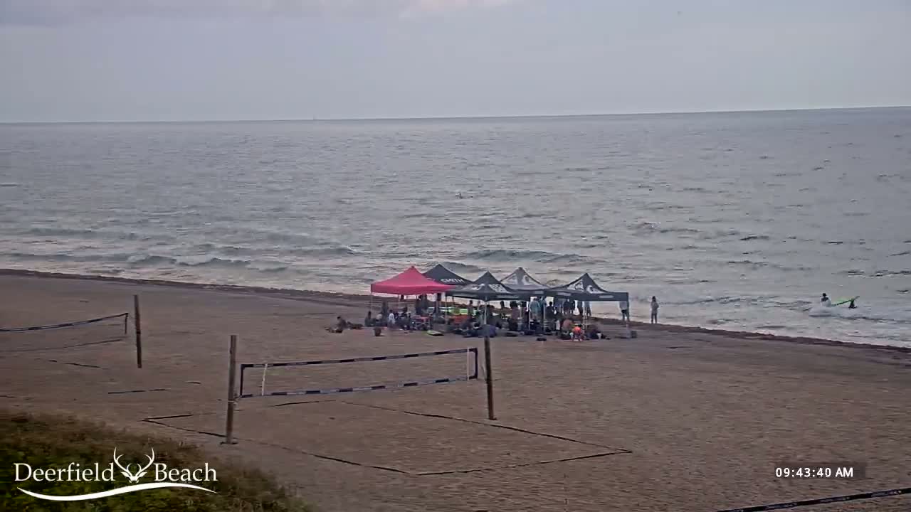 A sandy beach with several canopies, a group of people, and volleyball nets, overlooks a calm ocean with small waves under a cloudy sky.