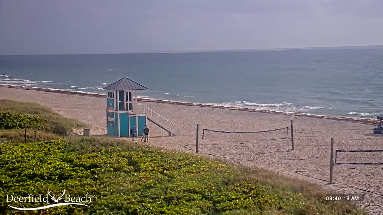 A sandy beach with a lifeguard stand, volleyball nets, and a few people, under a mostly cloudy sky and with gently rolling waves in the ocean.