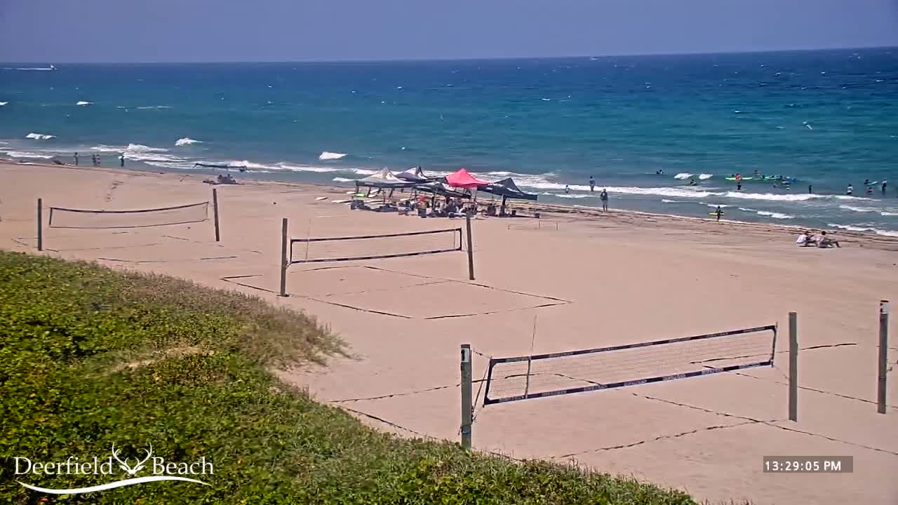 A sandy beach with several volleyball courts, people enjoying the water and sun, and a calm, teal ocean under a bright sunny sky.