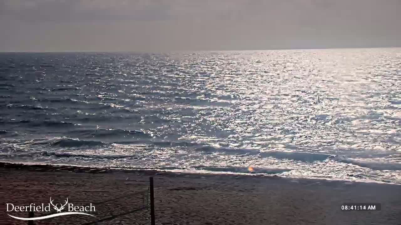 A sandy beach meets a moderately wavy ocean under a bright, somewhat overcast sky.