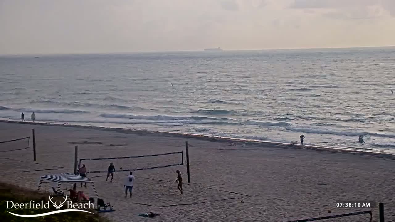 A sandy beach with people playing volleyball, a calm ocean with small waves, and a hazy sky.