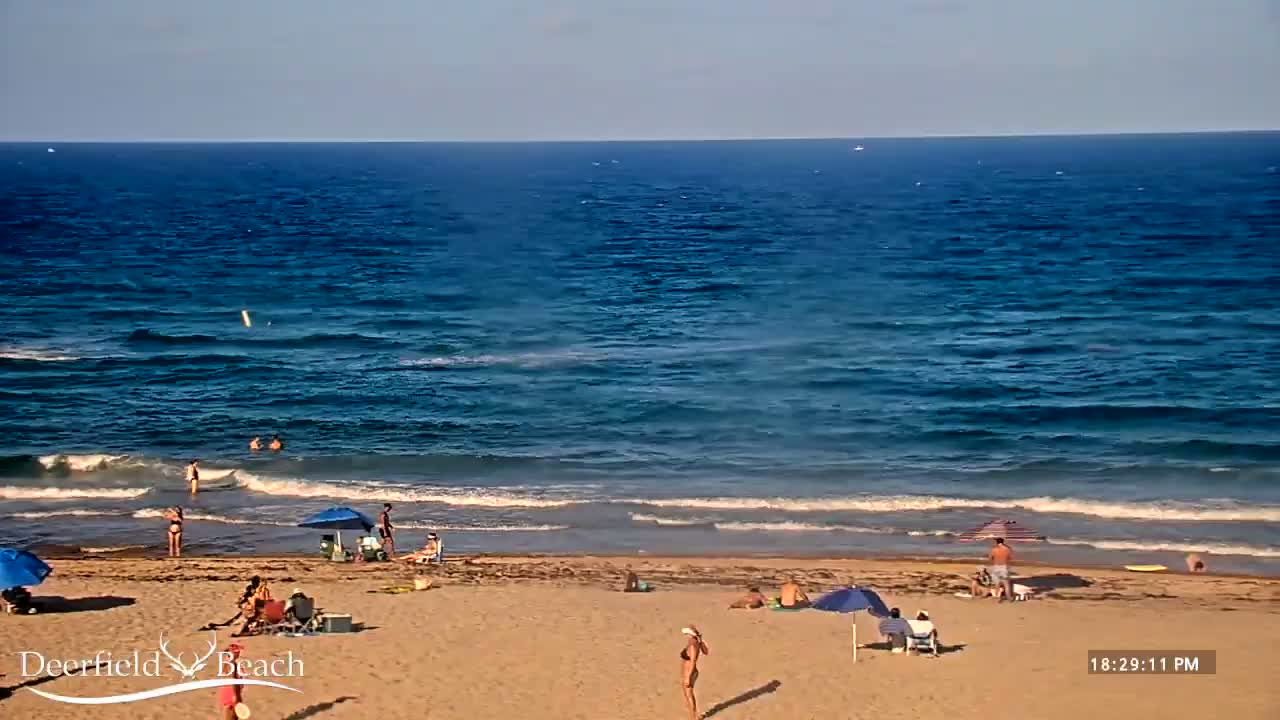 A sandy beach with people under umbrellas and a calm ocean with small waves under a partly cloudy sky.