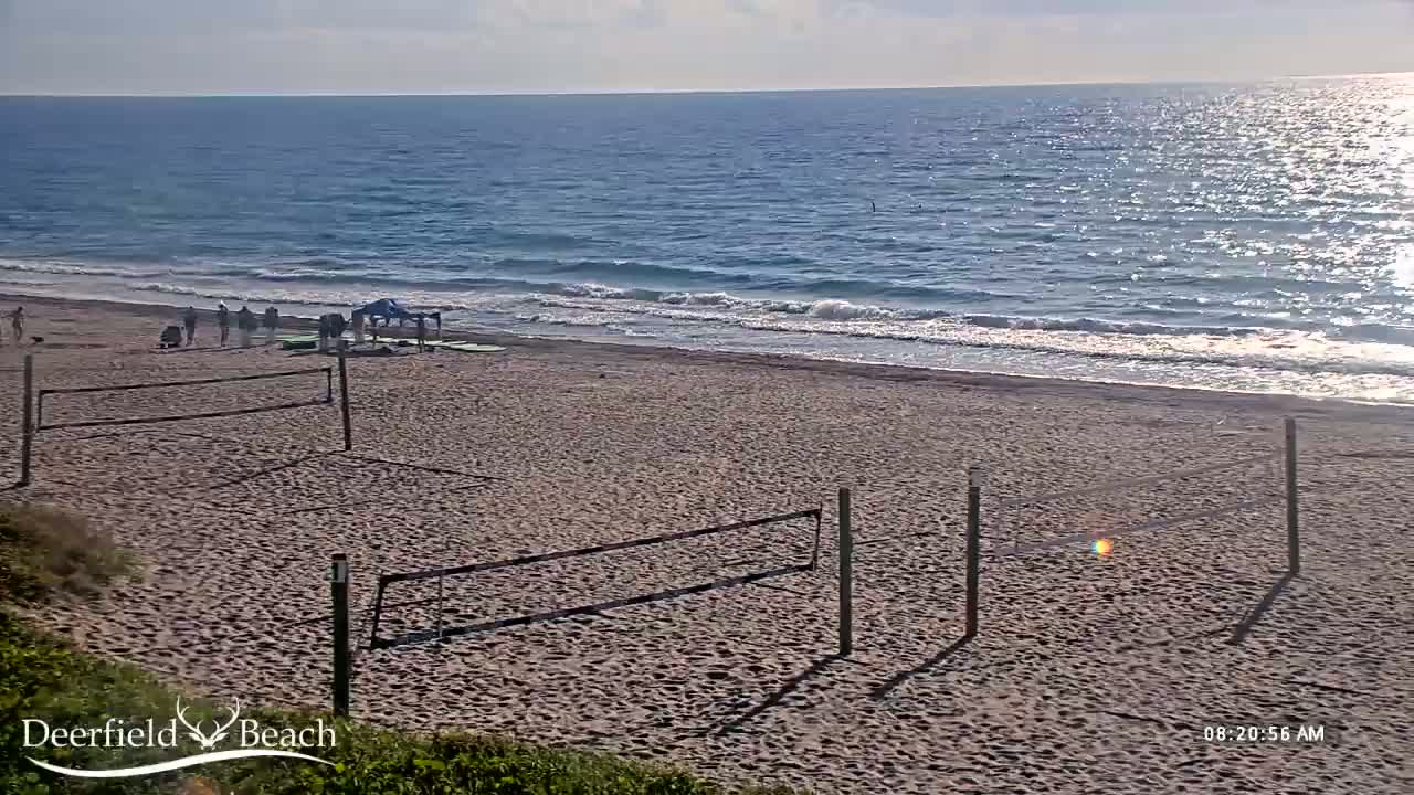 A sandy beach with volleyball nets and a few people is visible under a partly cloudy sky, with a calm ocean in the background.