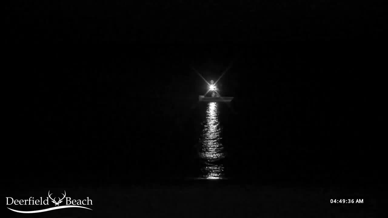A small boat with a bright, star-like light shining from its top casts a shimmering reflection on calm water under a clear, dark night sky.