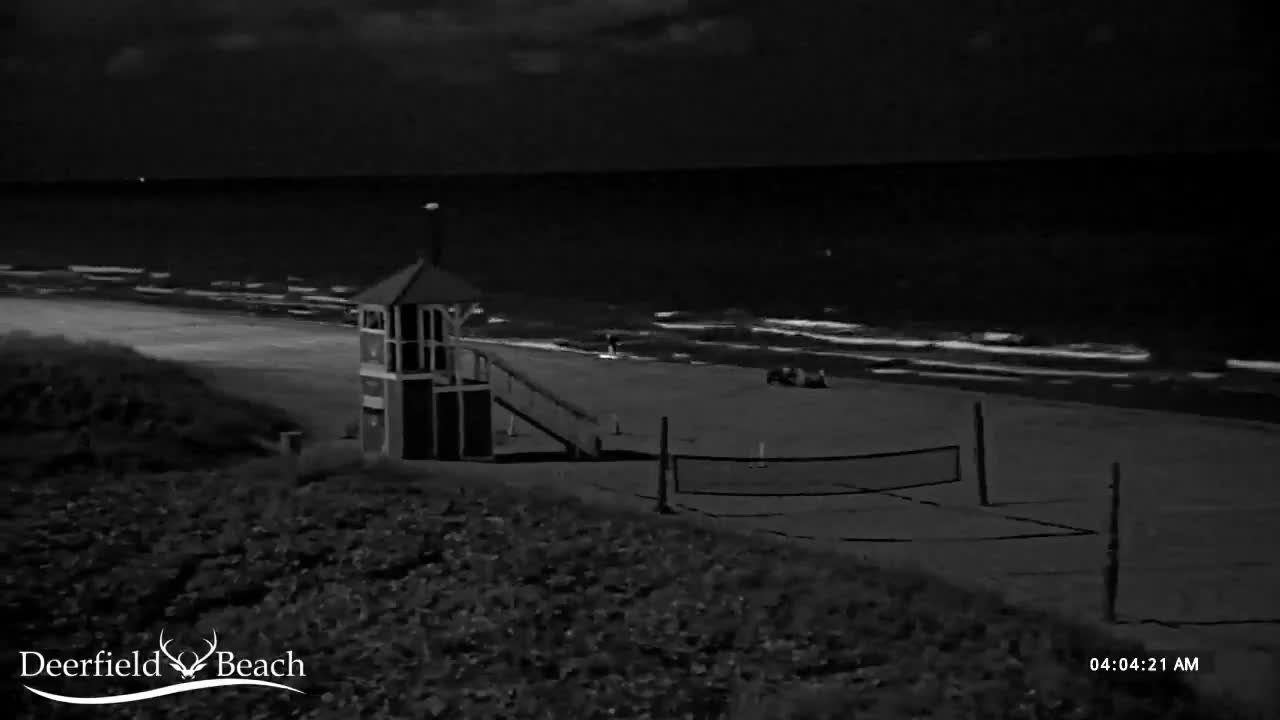 A dark, nighttime view captures a beach with a prominent lifeguard stand, a volleyball net, and gentle waves breaking on the shore under a clear or sparsely cloudy sky.