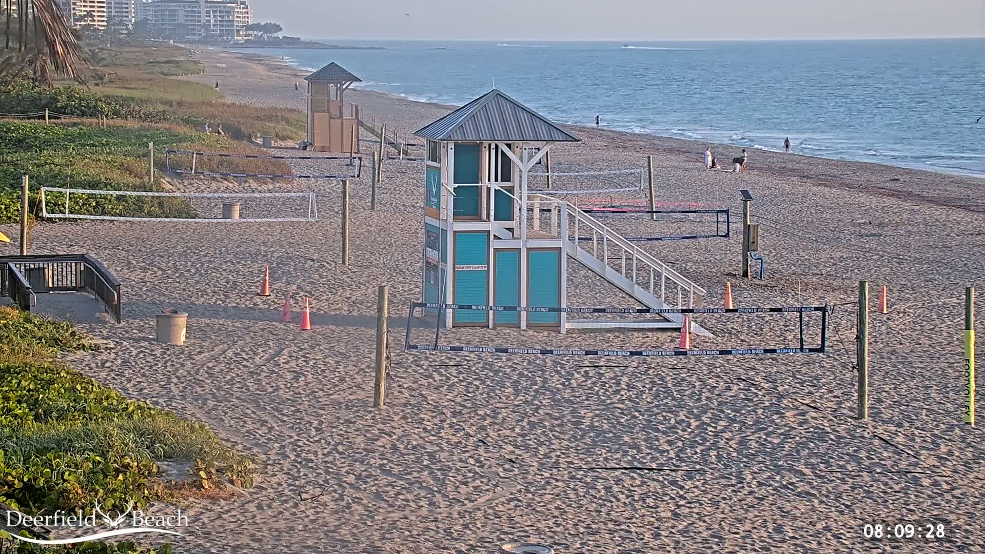 A sandy beach with volleyball courts and a lifeguard stand, under a mostly clear sky, overlooks a calm ocean.