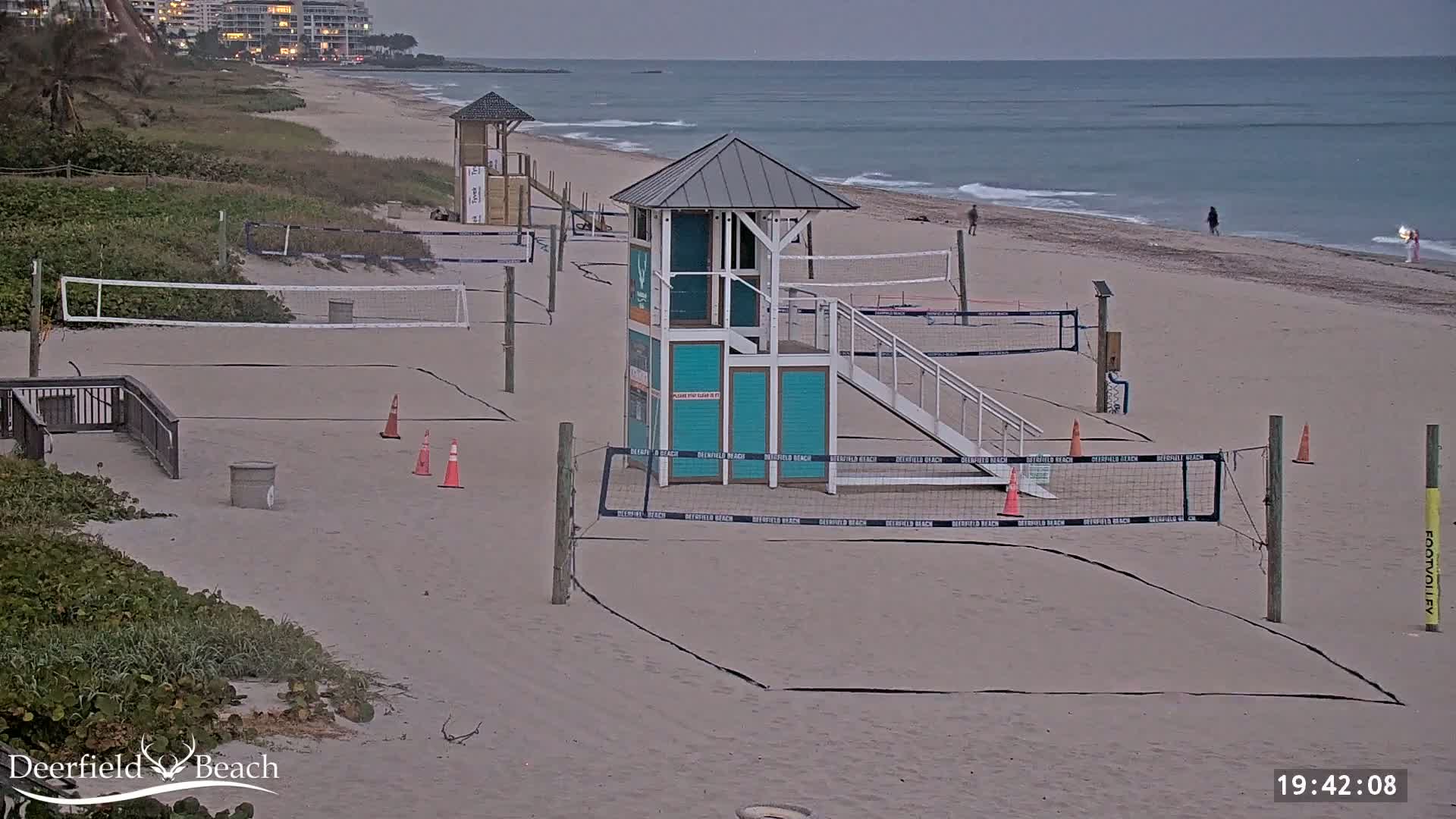 Several volleyball courts and lifeguard stands are situated on a sandy beach under overcast skies, with a calm ocean in the background.