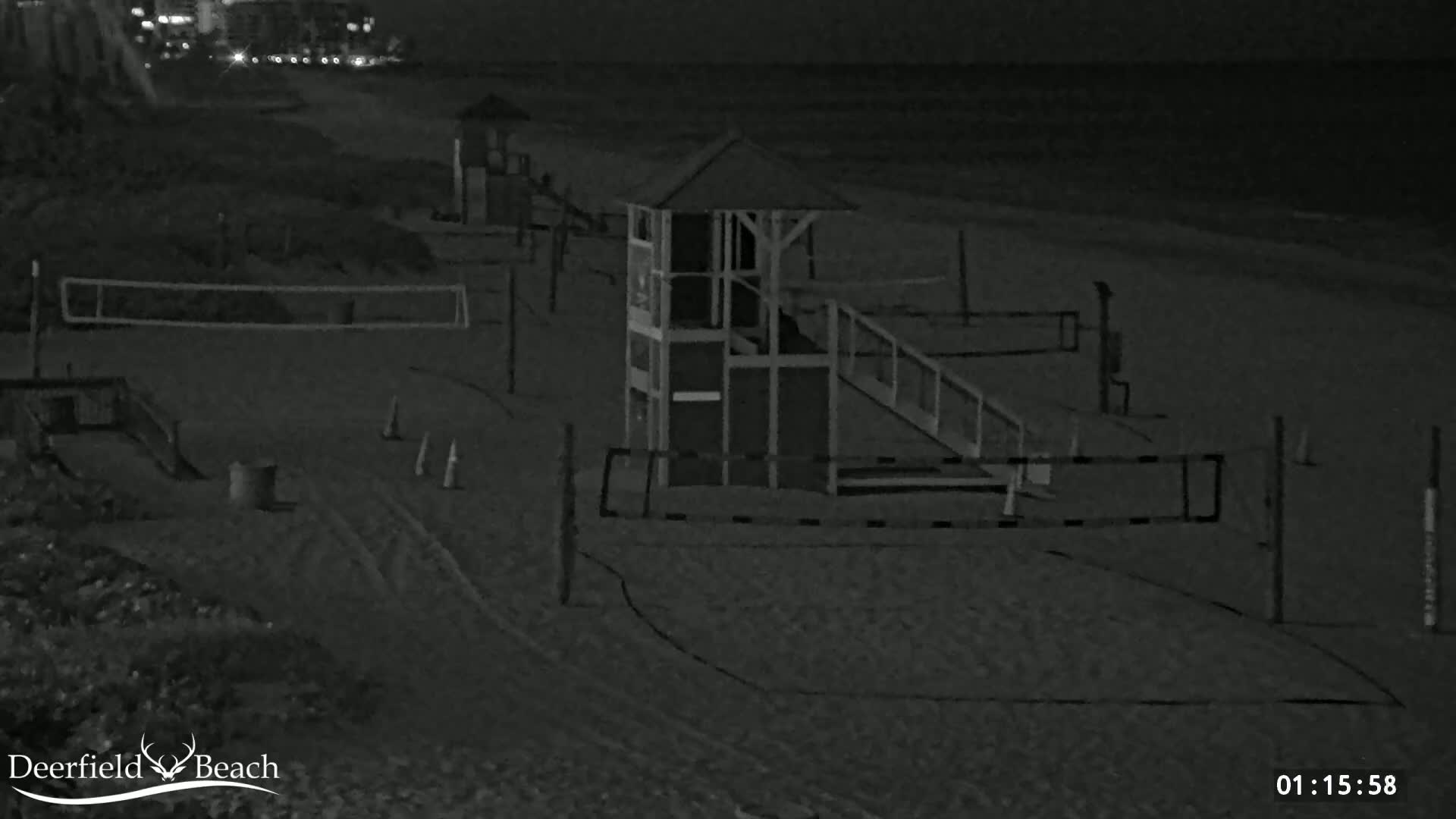 A nighttime, black and white image shows a mostly empty beach with volleyball nets, a lifeguard stand, and some other beach equipment.