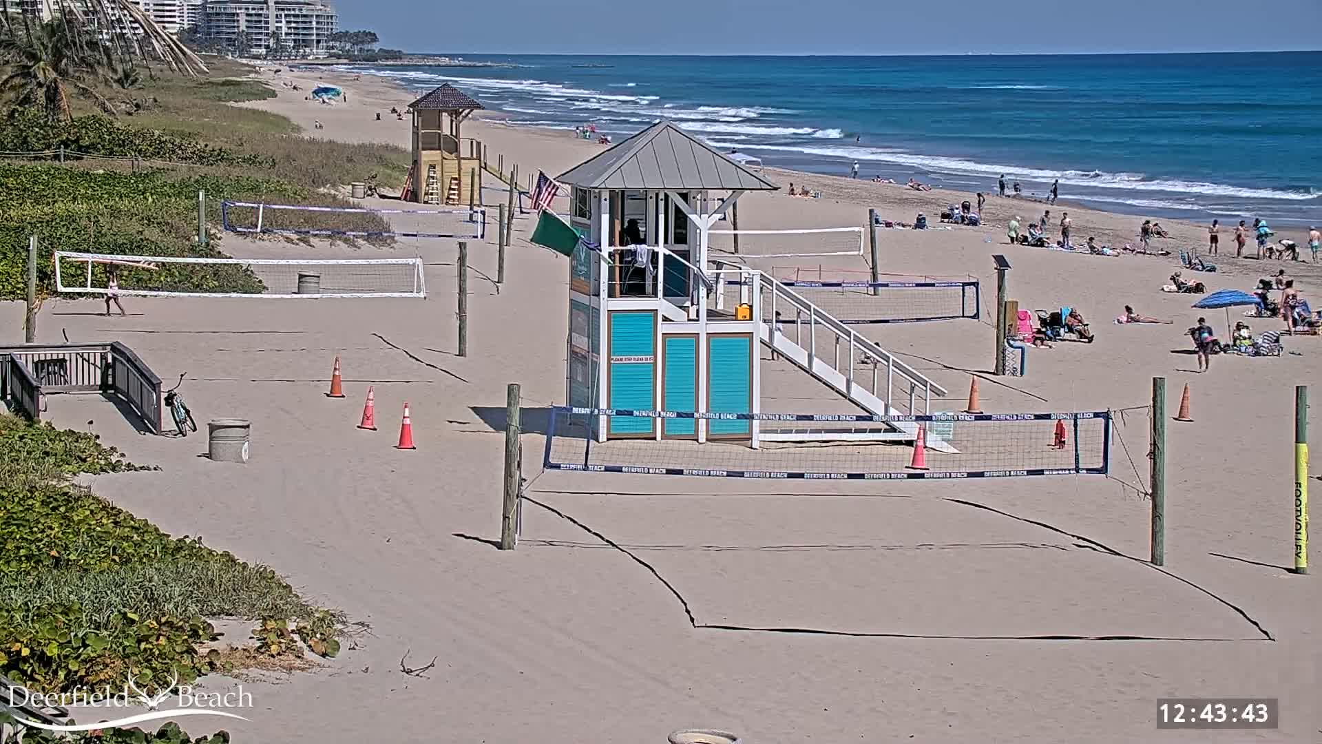 A sandy beach with volleyball courts, lifeguard stands, and people relaxing under a sunny sky and calm ocean.