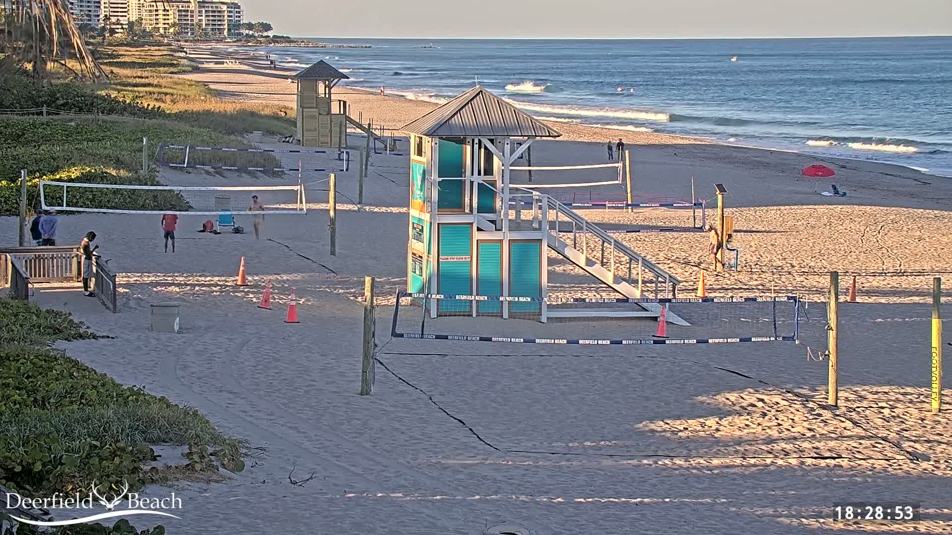 A sandy beach with volleyball courts, lifeguard stands, and a few people is visible under sunny skies and calm ocean waves.