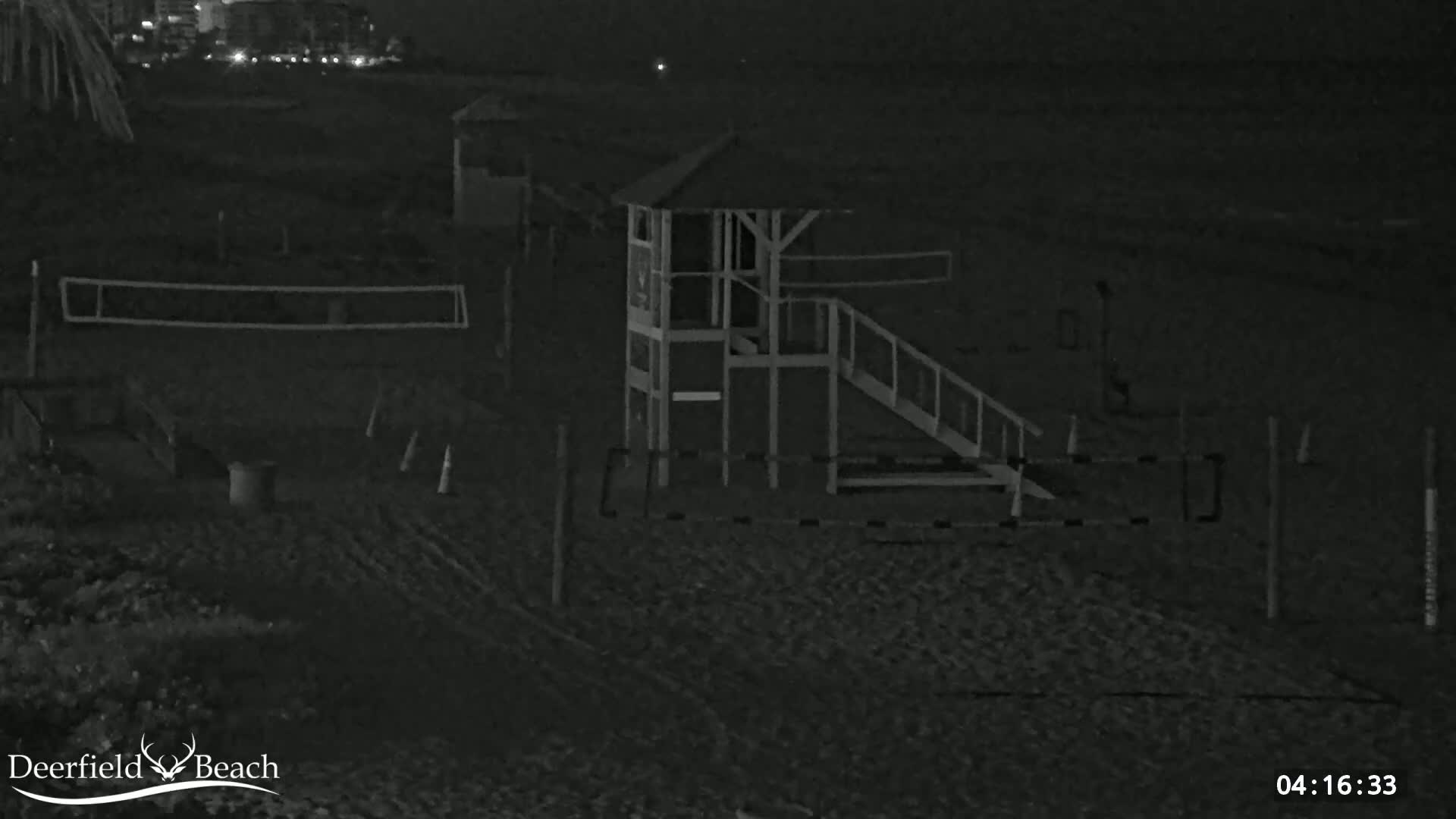 A dark, nighttime beach scene shows a volleyball net, a lifeguard stand with a ramp, and other beach equipment under a dark sky.