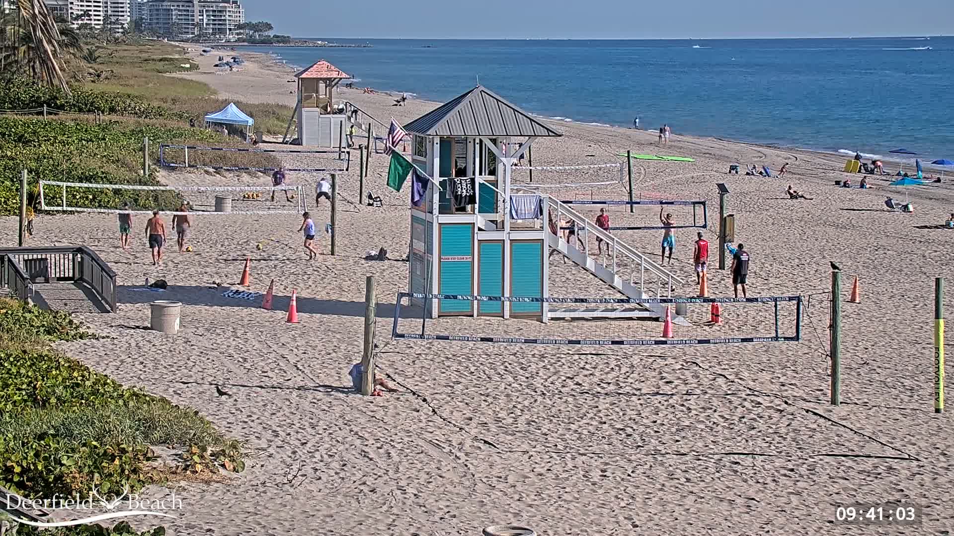 A sandy beach on a sunny day features several volleyball courts with people playing, a lifeguard stand, and the ocean in the background.