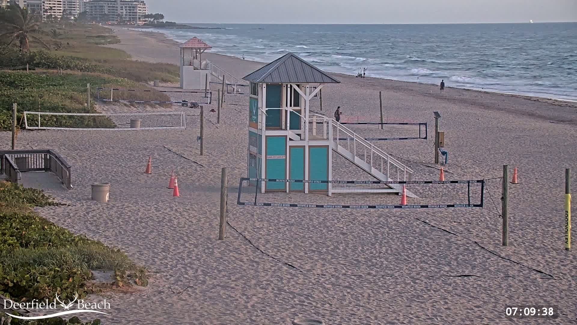 A sandy beach with volleyball nets, lifeguard stands, and a calm ocean under a mostly clear sky.