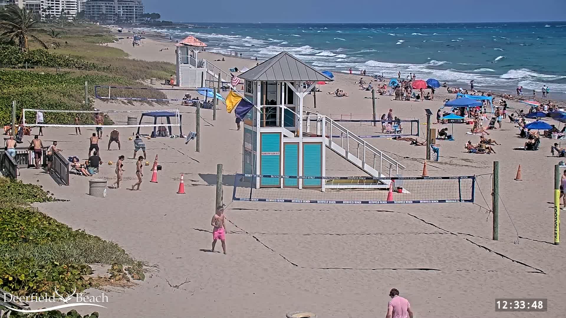 A sandy beach with volleyball courts, lifeguard stands, and many people enjoying a sunny day with a somewhat wavy ocean in the background.