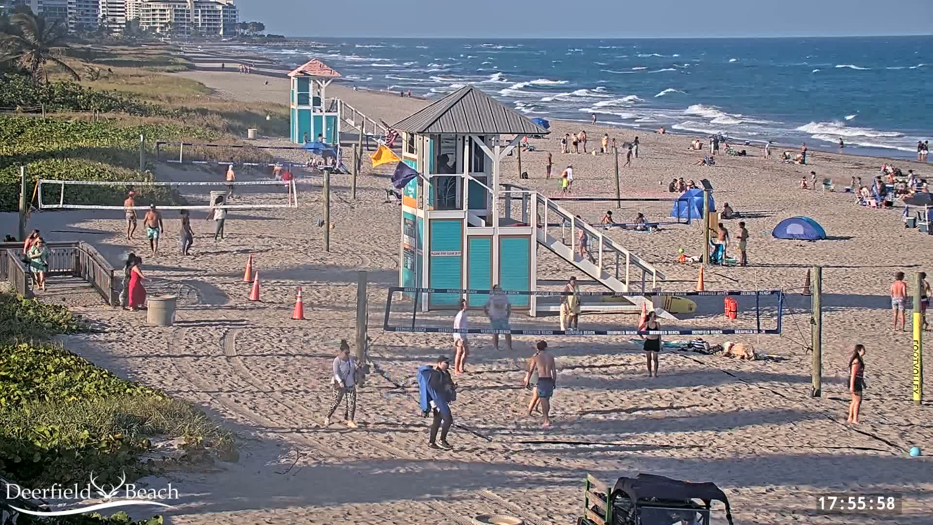 A sandy beach with multiple volleyball courts, lifeguard stands, and numerous people enjoying a sunny day with somewhat wavy ocean waters.