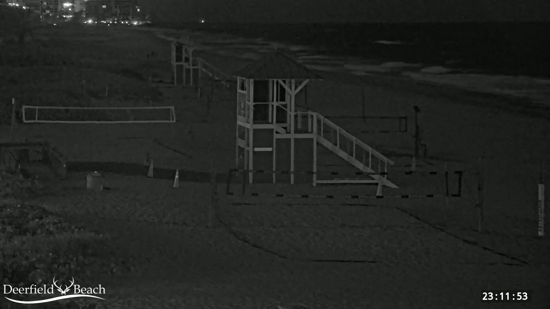 A nighttime beach scene shows a lifeguard stand with a ramp, a volleyball net, and calm ocean waves.