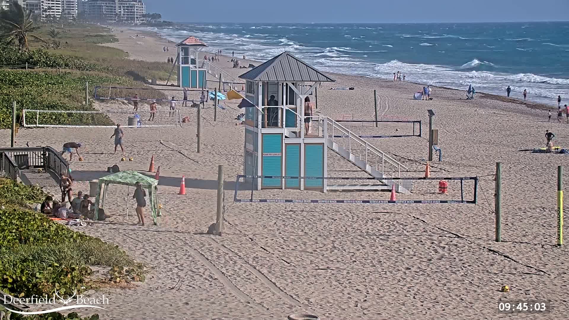 A sandy beach with several volleyball nets, lifeguard stands, and people playing sports and relaxing under a partly cloudy sky, with a windy ocean in the background.