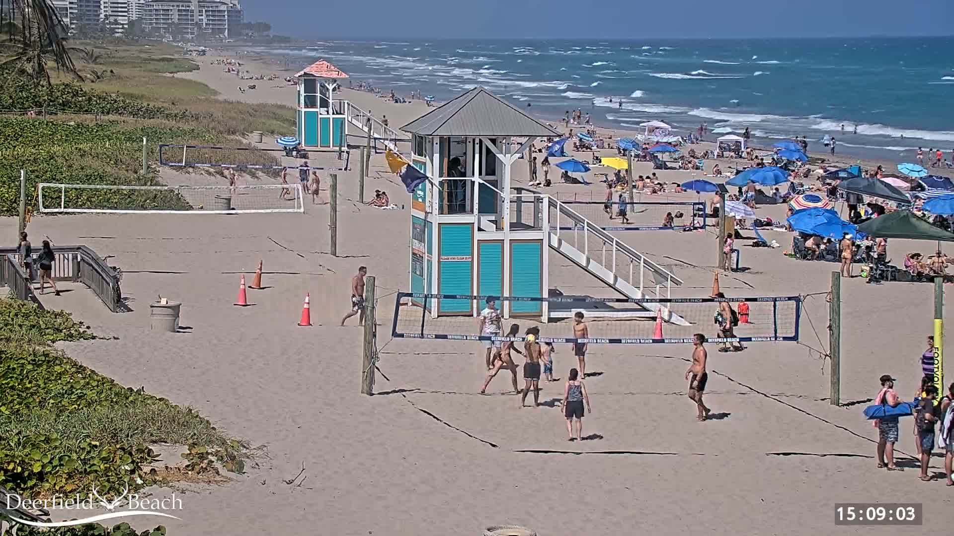 A sandy beach with volleyball courts, lifeguard stands, and many people enjoying a sunny day with a wavy ocean in the background.