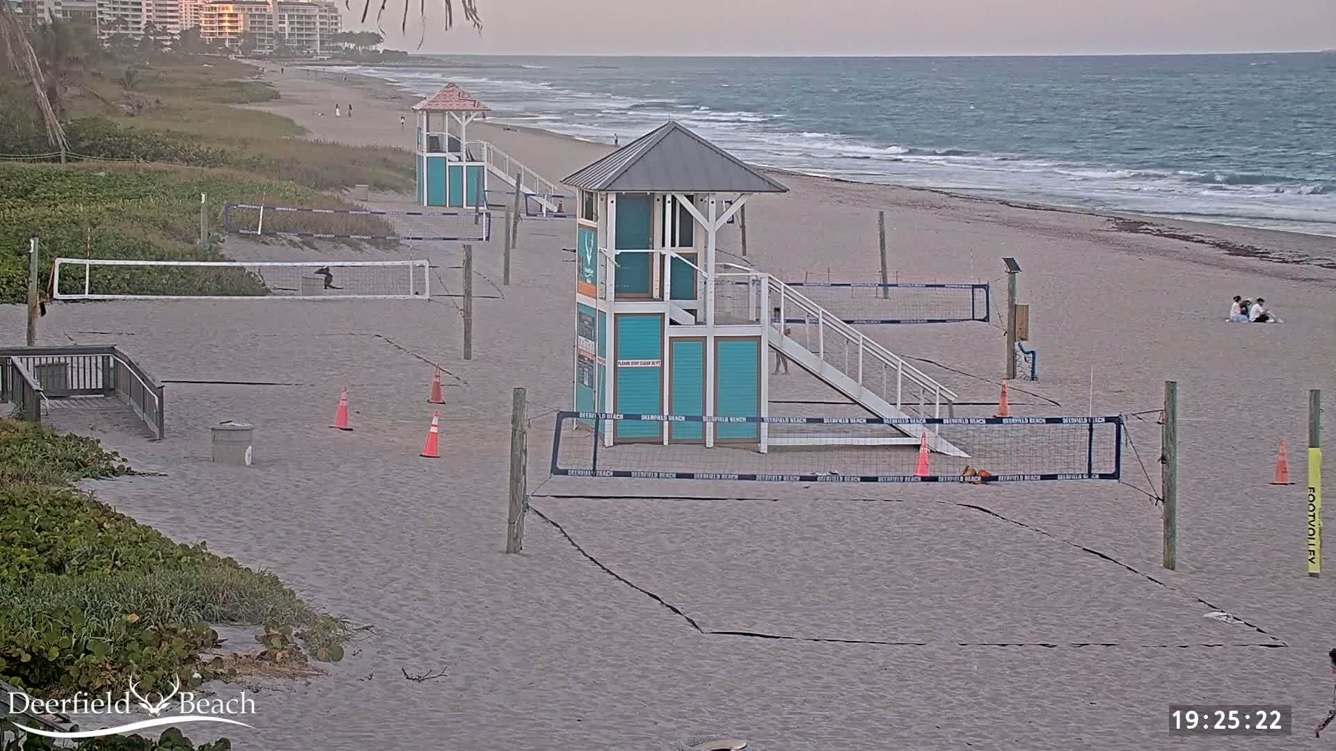 A sandy beach with several volleyball courts and lifeguard stands under an overcast sky, next to a calm ocean.
