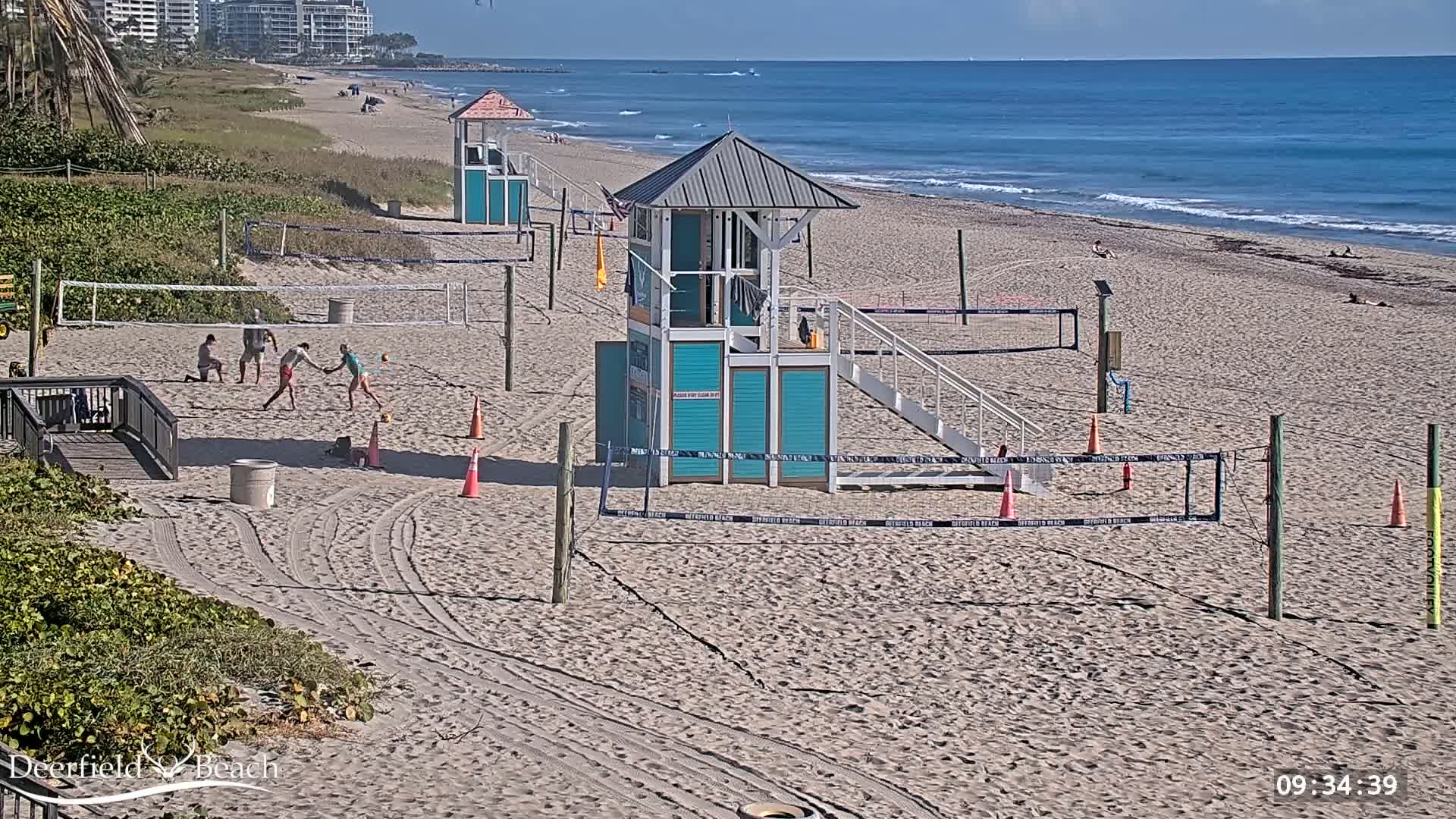 A sandy beach with several volleyball nets, lifeguard stands, and a few people playing volleyball under a clear, sunny sky.