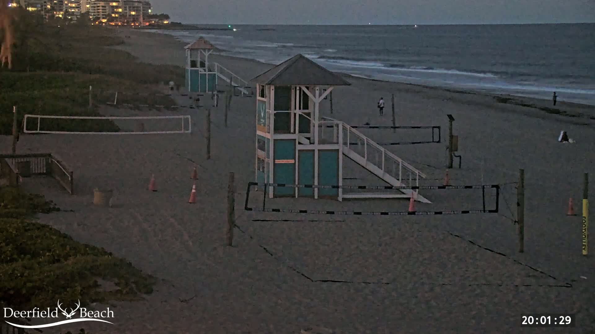 A sandy beach at dusk shows two lifeguard stands, volleyball nets, and a few people near the water's edge under a mostly clear sky.