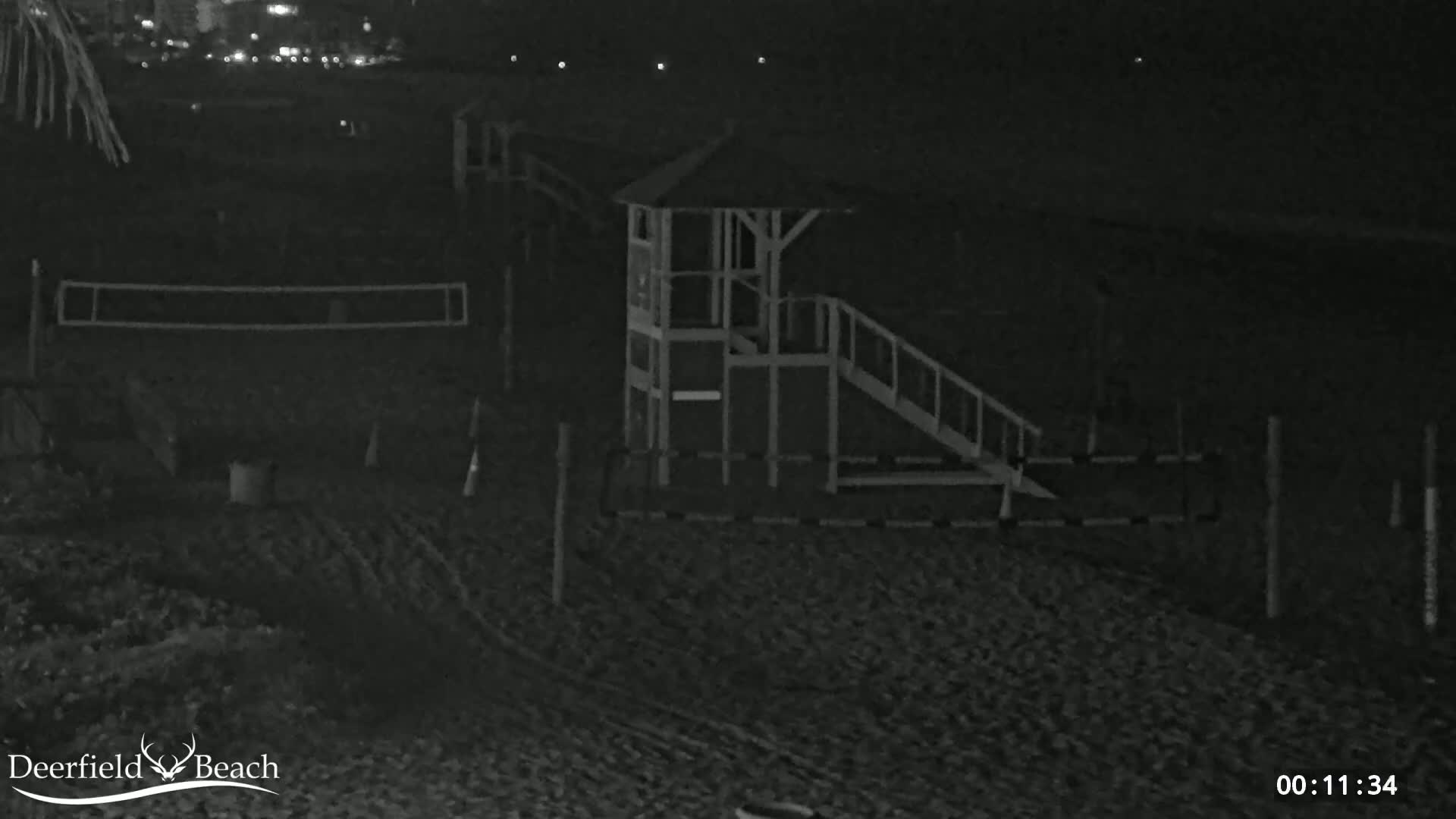 A nighttime, low-light image shows a sandy beach with a lifeguard stand, a volleyball net, and some fencing.