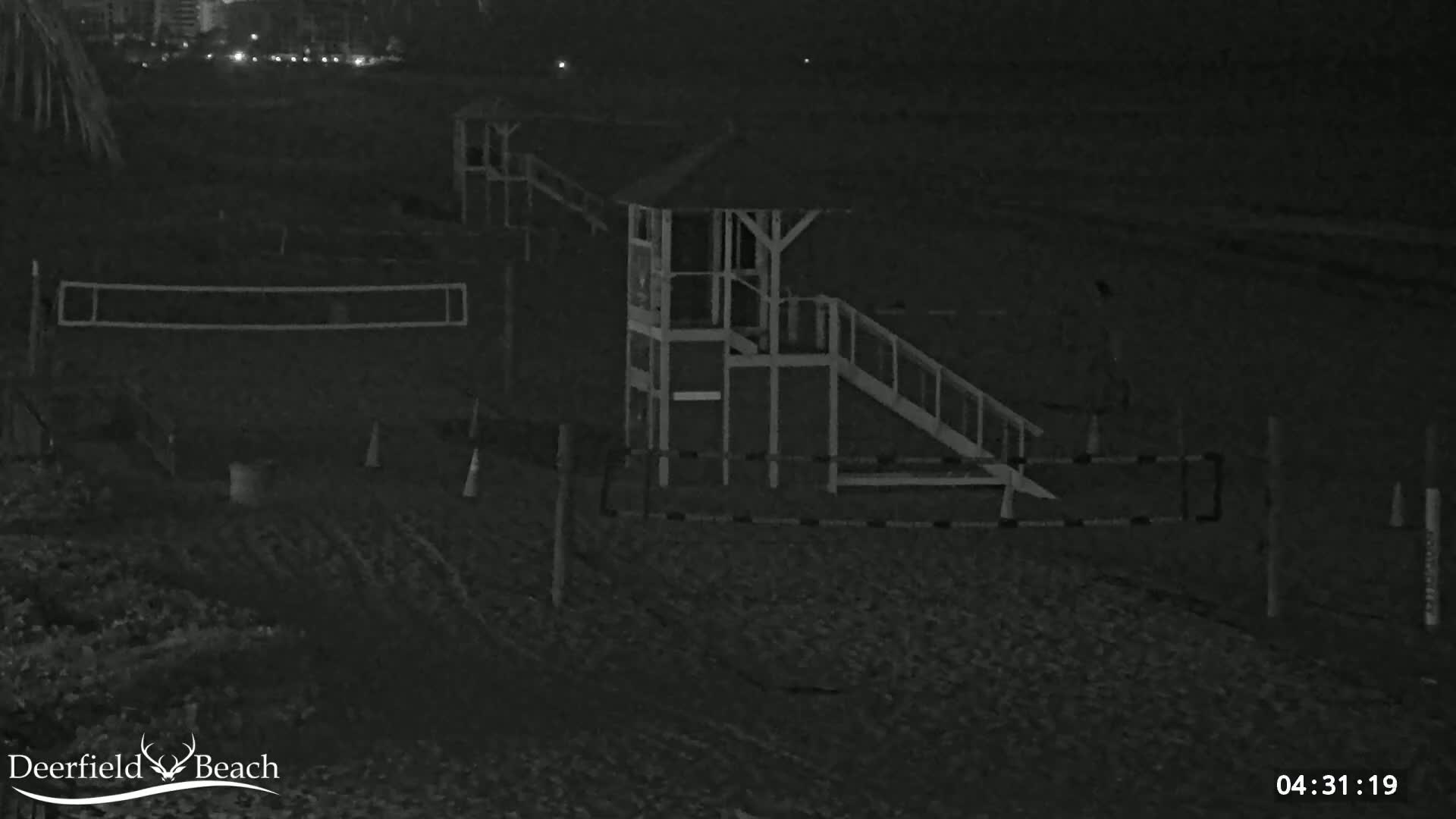 A nighttime, low-light image shows a beach volleyball court, a lifeguard stand with a ramp, and a fence on a dark, sandy beach.