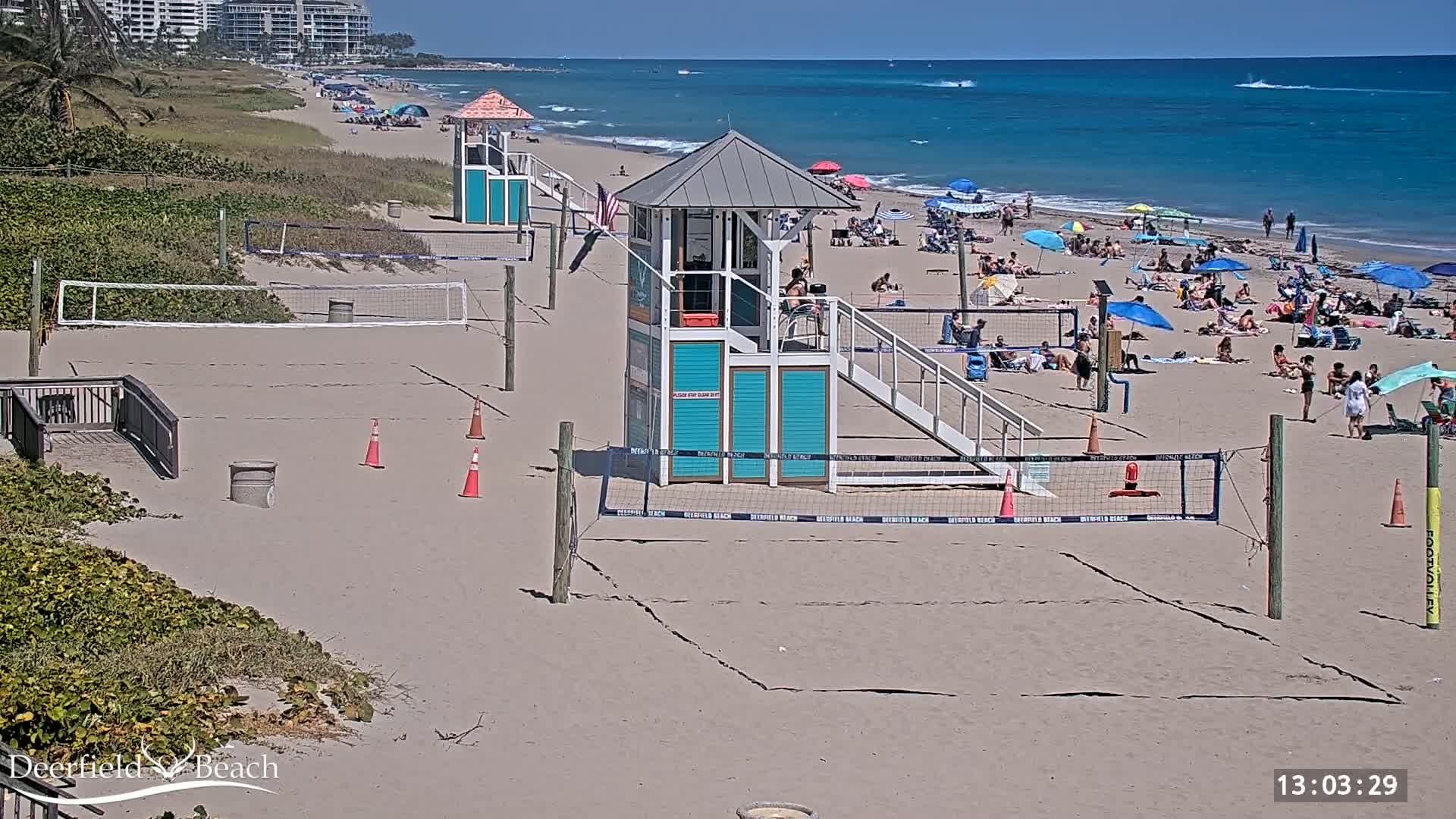 A sandy beach with volleyball courts, lifeguard stands, and numerous people enjoying a sunny day.