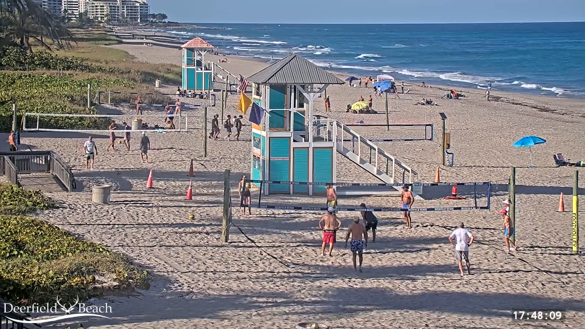 A sandy beach with multiple volleyball games in progress, lifeguard stands, and people sunbathing, all under a sunny sky and with a calm ocean in the background.