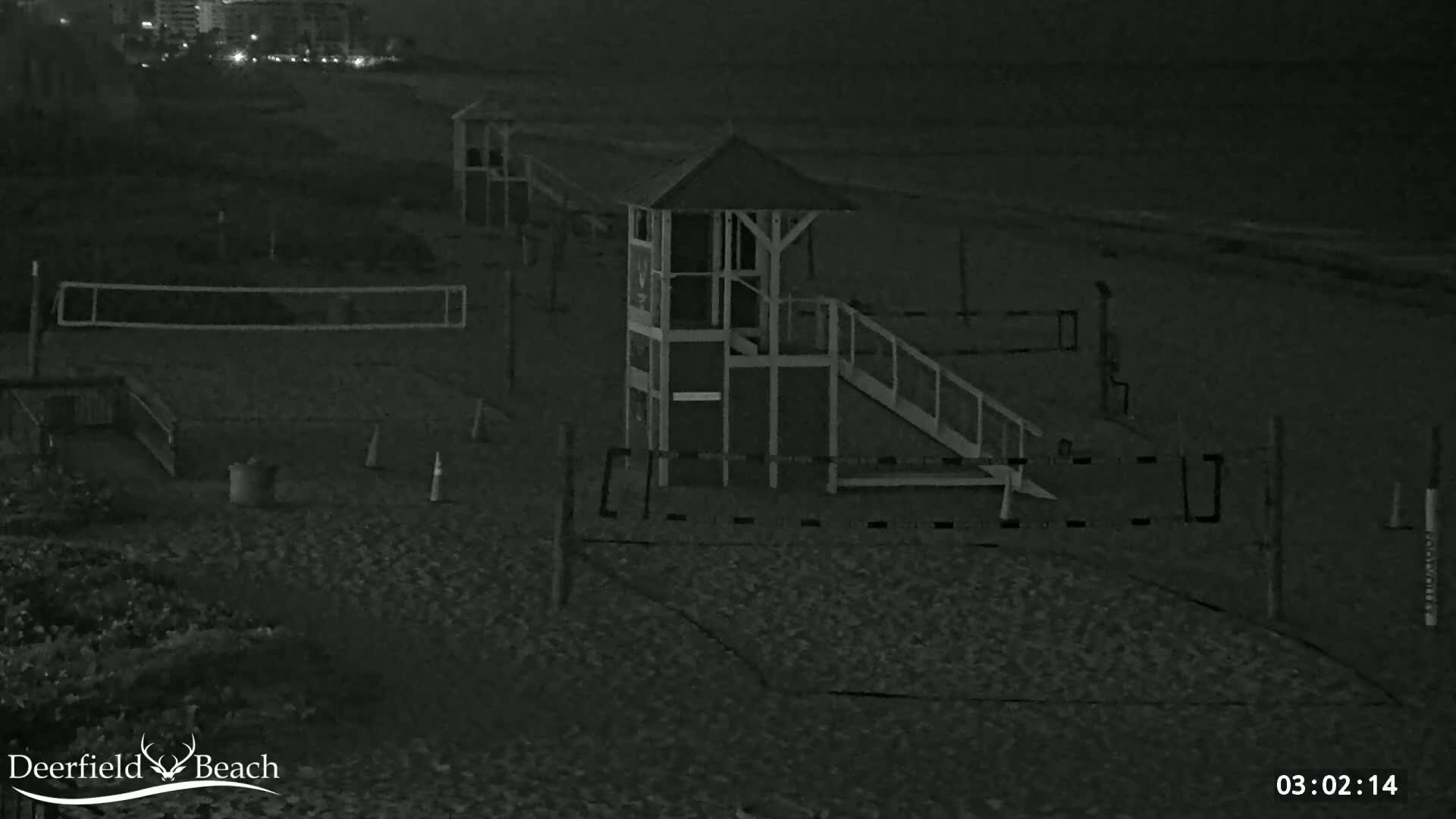 A nighttime, black-and-white view of a beach volleyball court with a lifeguard stand and a dark ocean in the background.