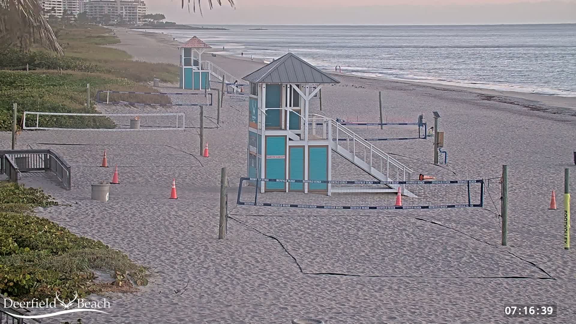 A sandy beach with volleyball courts, lifeguard stands, and calm ocean waves under an overcast sky.
