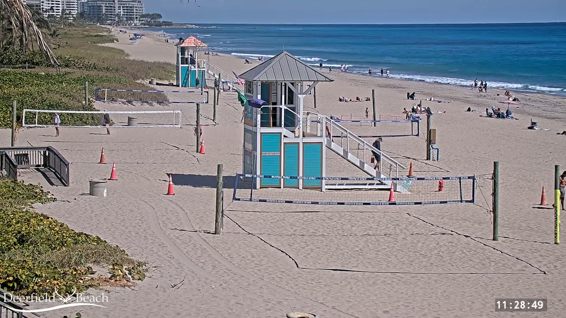 A sandy beach with volleyball courts, lifeguard stands, and scattered beachgoers on a sunny day with calm, light blue ocean waves.