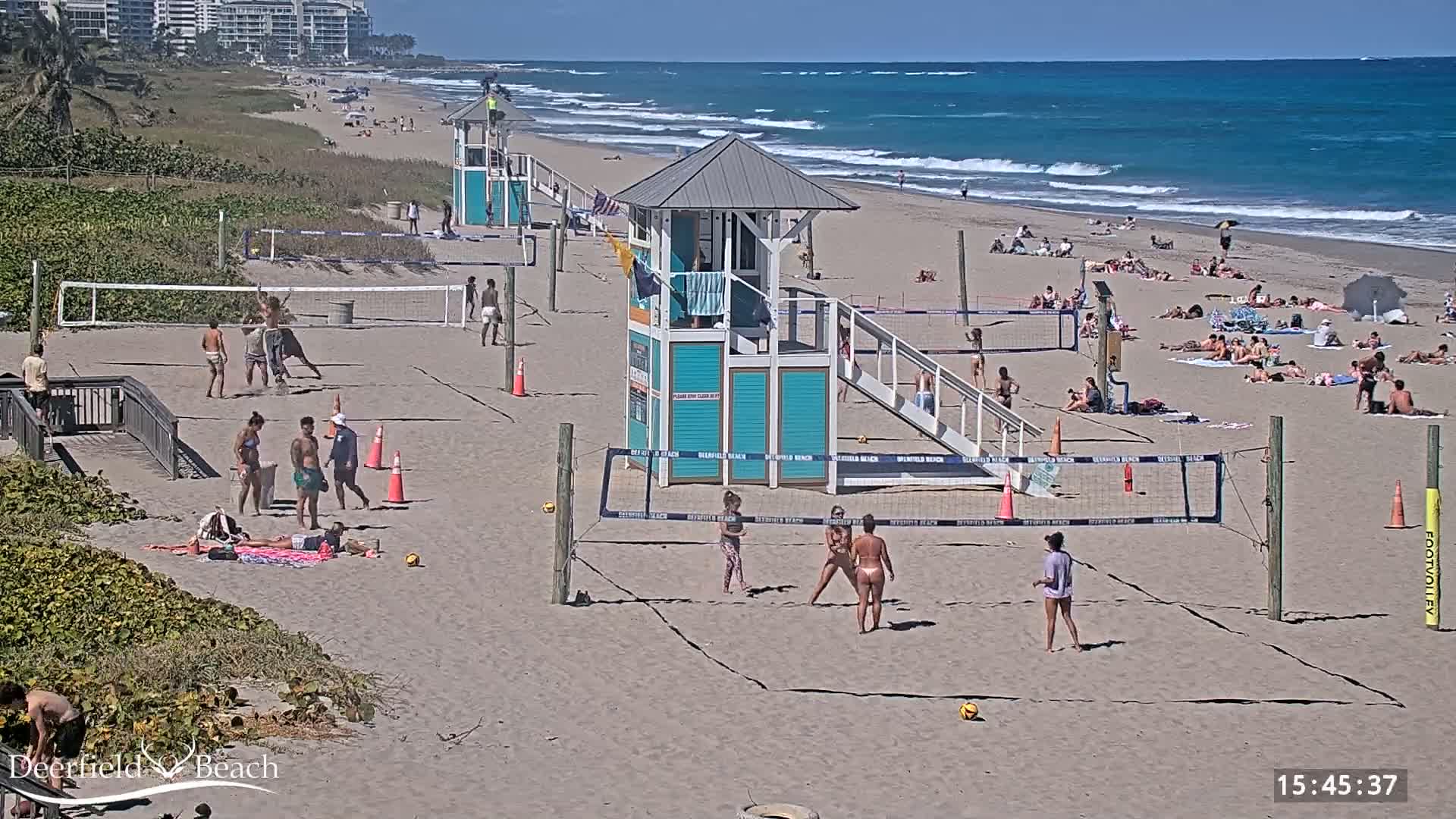 A sandy beach with several volleyball courts, lifeguard stands, and many people sunbathing and playing volleyball on a sunny day with ocean waves visible in the background.