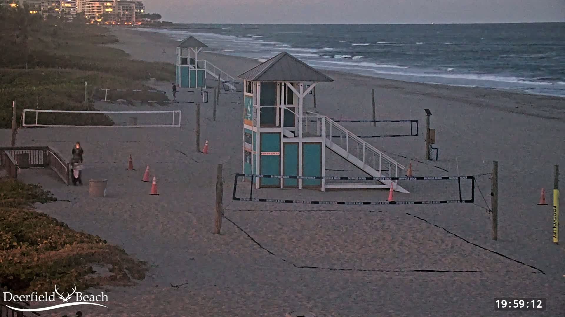 A sandy beach at dusk with two lifeguard stands, volleyball nets, and a few people walking near a boardwalk.