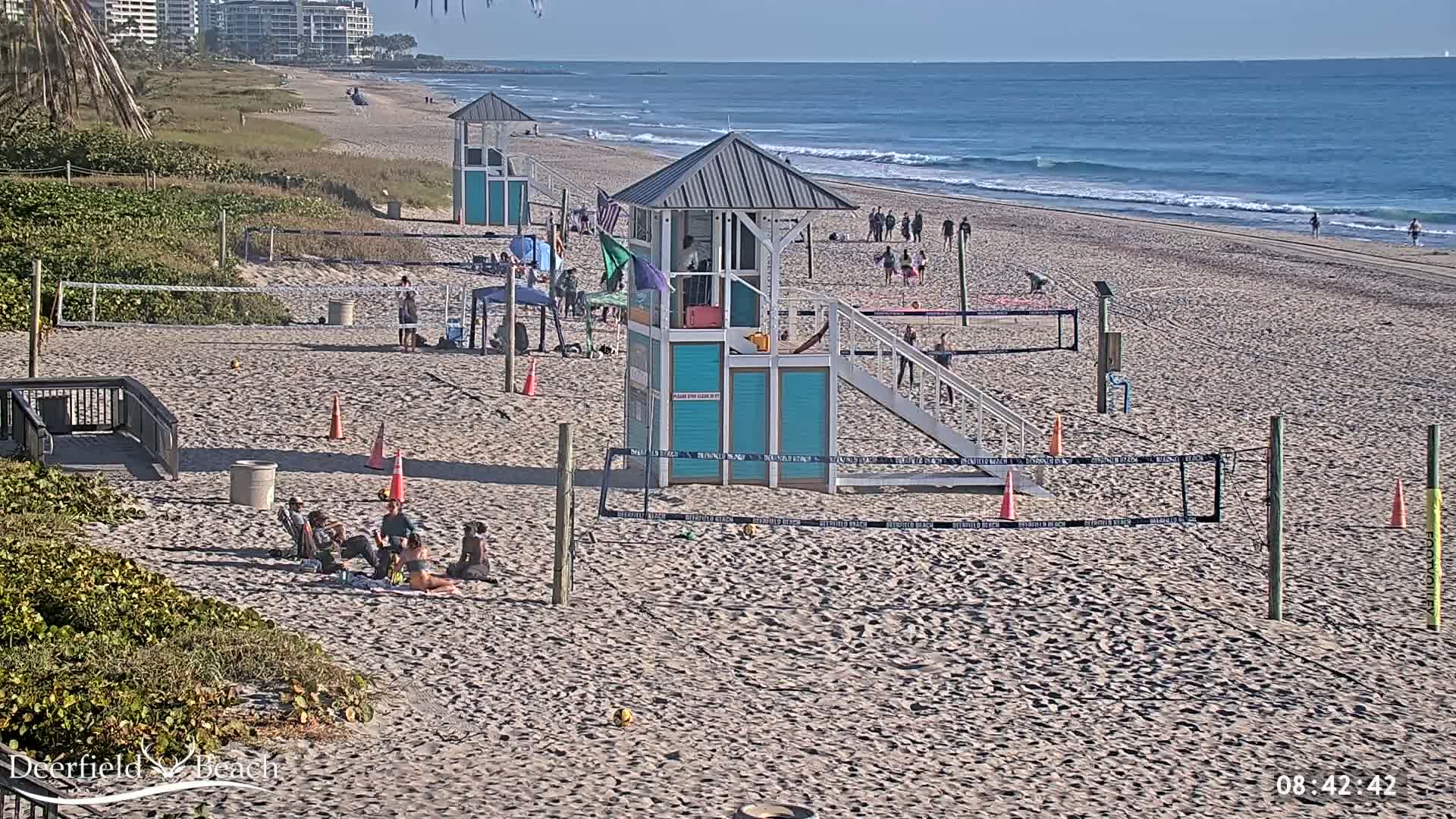 A sandy beach with calm ocean waves, several volleyball nets, lifeguard stands, and a few people scattered across the beach on a sunny day.