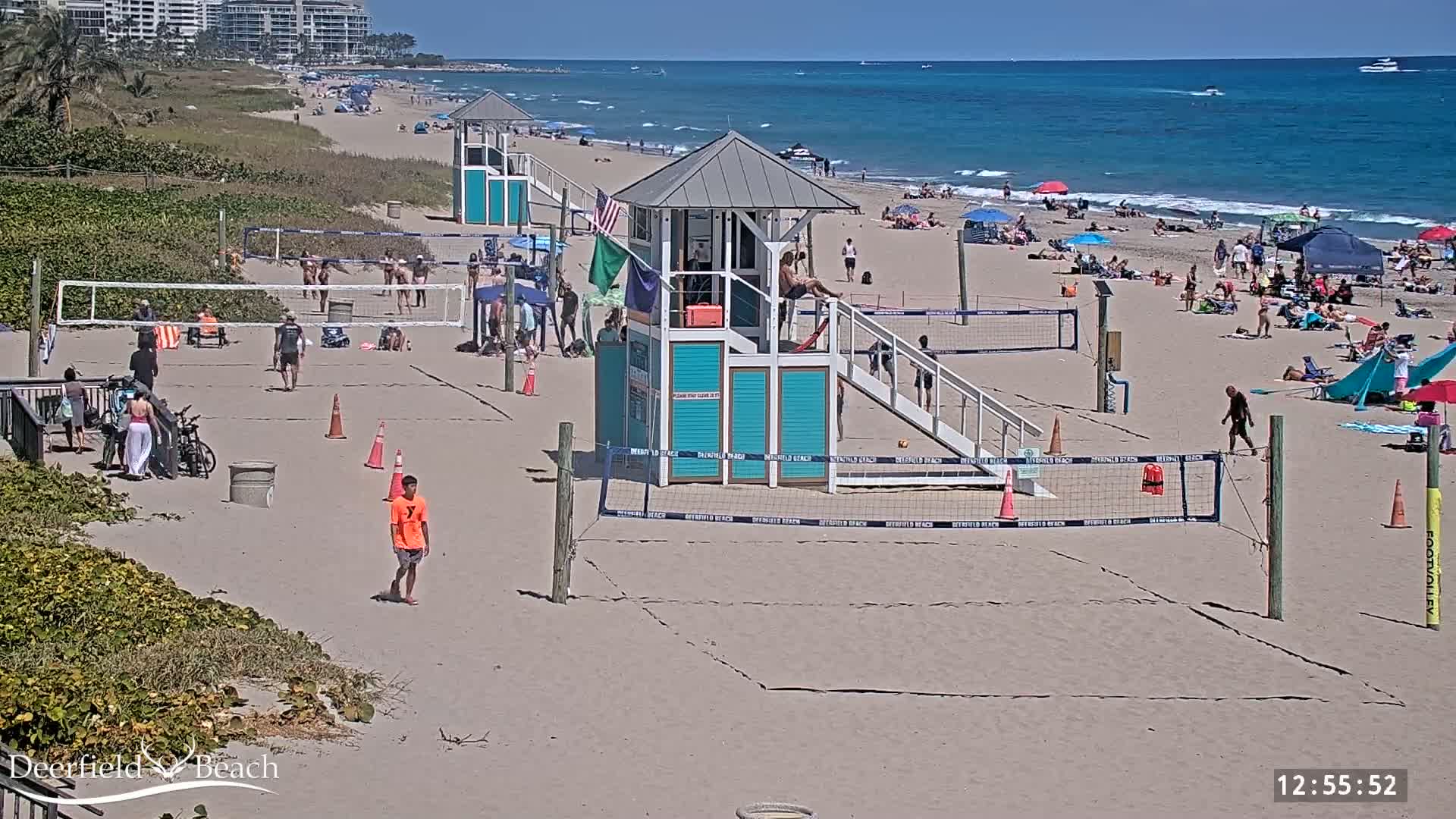A sunny beach scene shows several volleyball courts with people playing and relaxing on the sand, a lifeguard stand, and the ocean in the background.
