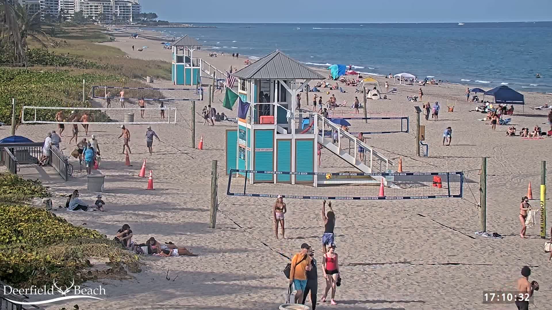 A sandy beach with several volleyball courts, lifeguard stands, and many people enjoying a sunny day.