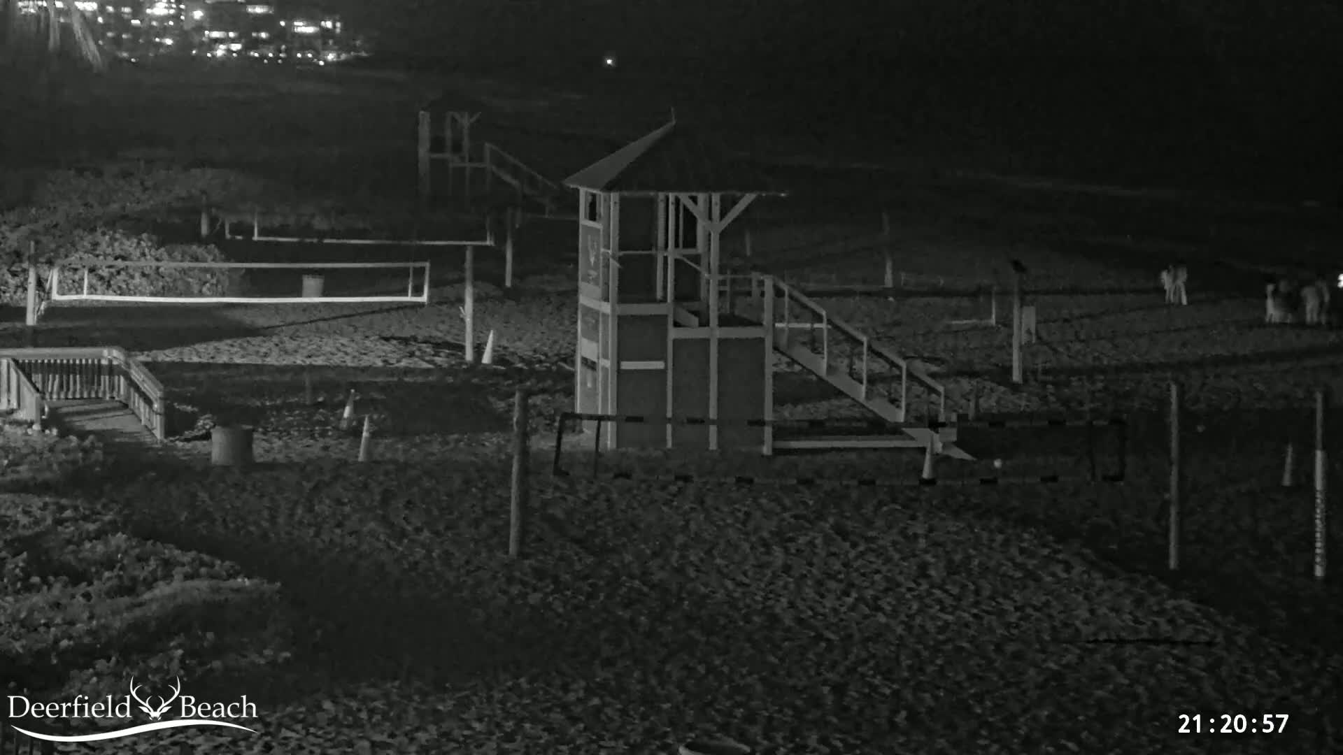 A nighttime, black and white image of a sandy beach with volleyball nets, a lifeguard stand, and a few people visible in the distance under dark skies.