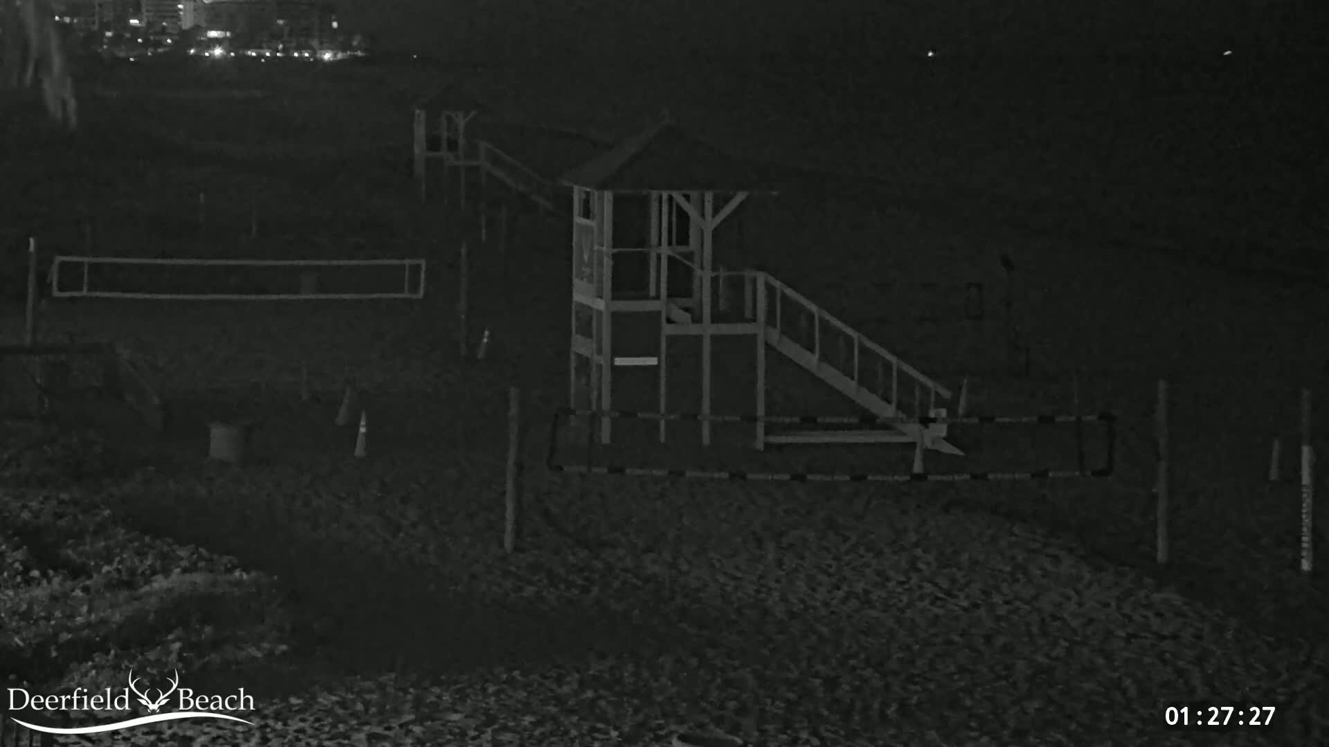 A nighttime, low-light image shows a beach with a lifeguard stand, volleyball net, and other beach equipment.