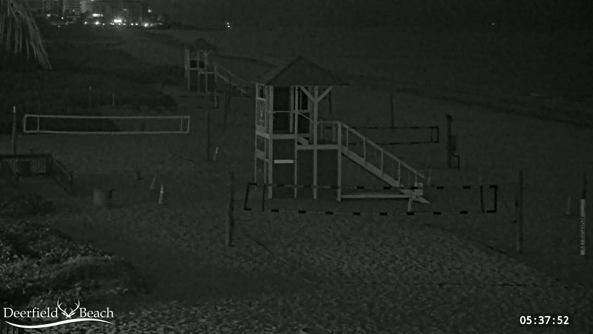 A nighttime, black-and-white image shows a sandy beach with volleyball nets, lifeguard stands, and other beach equipment under dark conditions.
