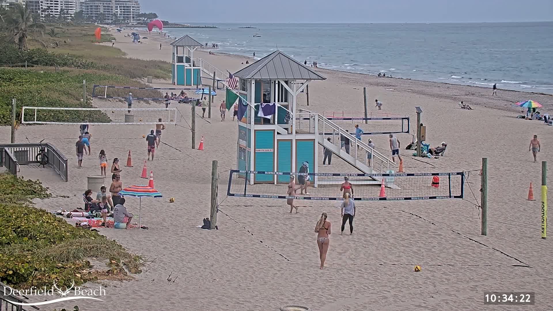 A sandy beach with multiple volleyball courts, lifeguard stands, and people playing volleyball and relaxing under umbrellas on an overcast day.