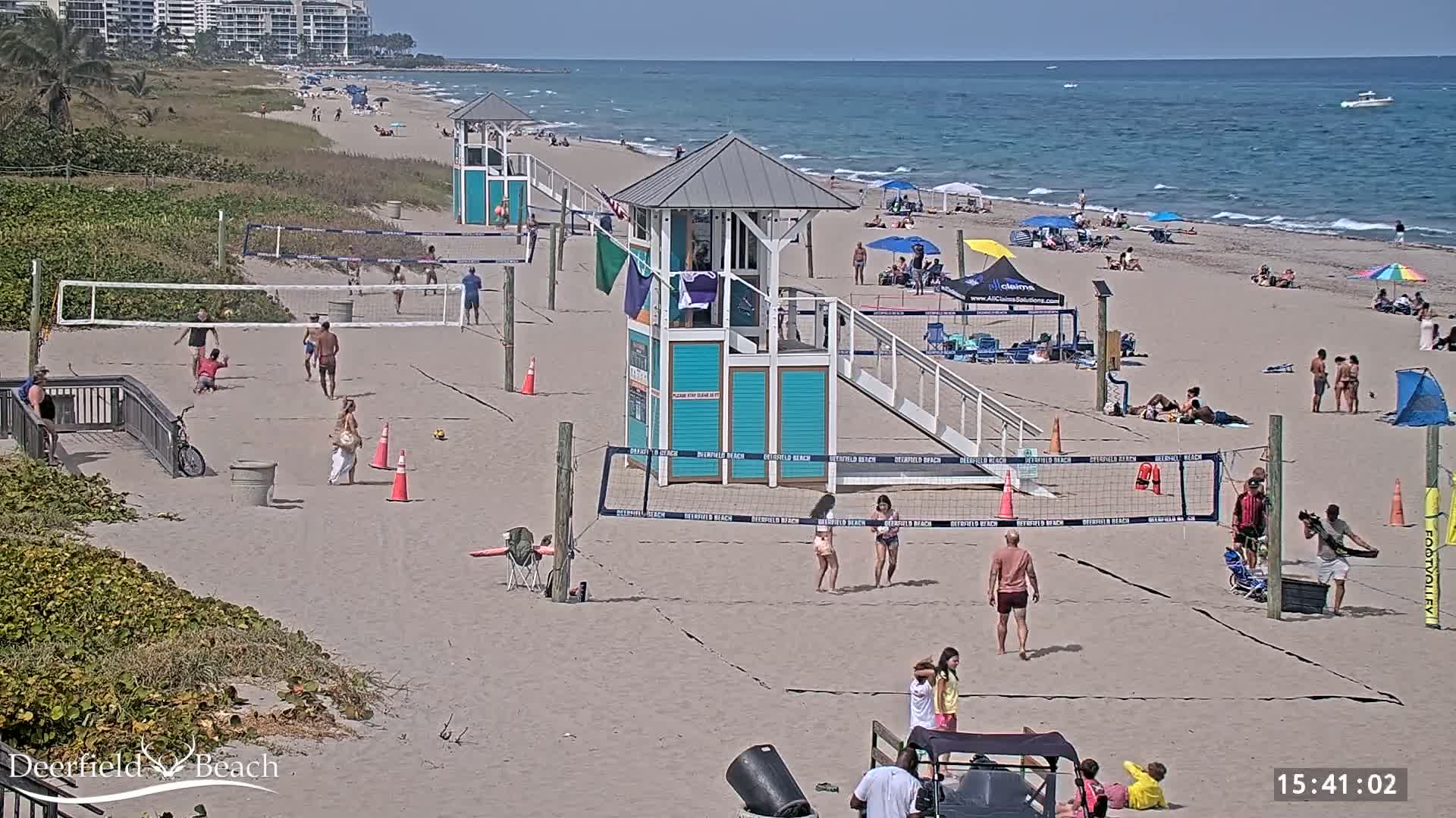 A sandy beach with multiple volleyball courts, lifeguard stands, and several people playing volleyball and relaxing under umbrellas on a sunny day with a calm ocean in the background.