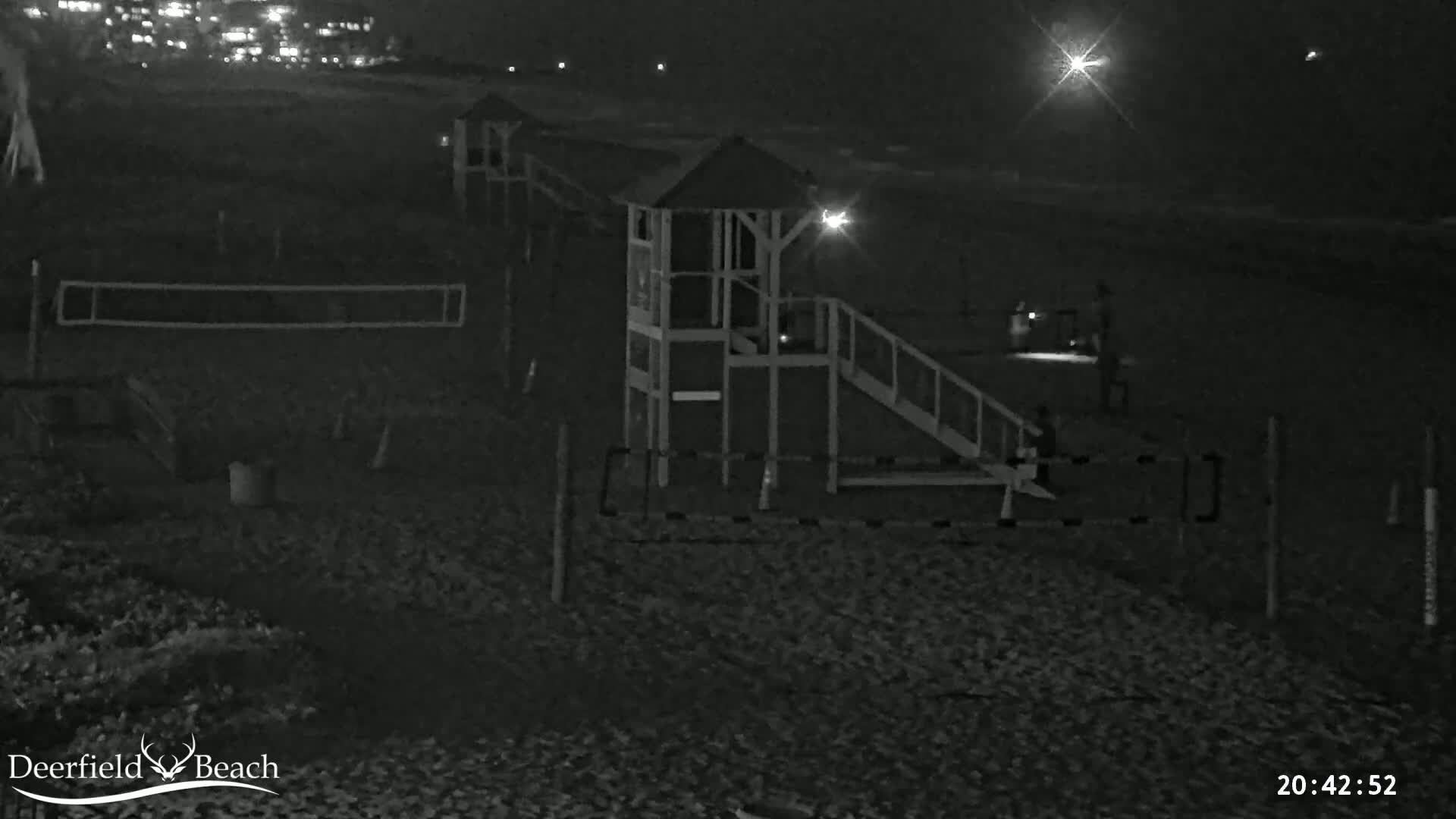 A nighttime, black-and-white image shows a beach with a lifeguard stand, volleyball net, and several people near the water under a bright light.