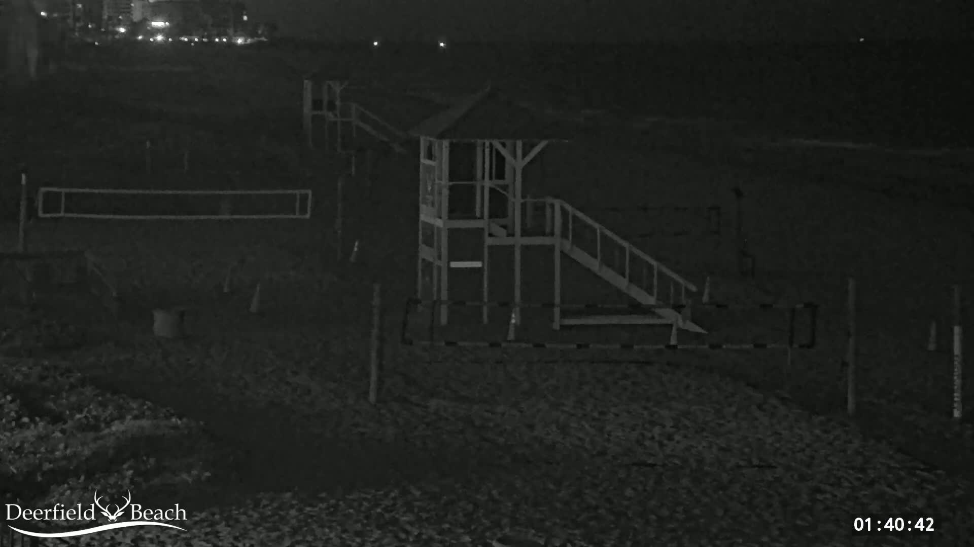 A nighttime view of a dark beach shows a lifeguard stand, volleyball nets, and a calm ocean in the background.