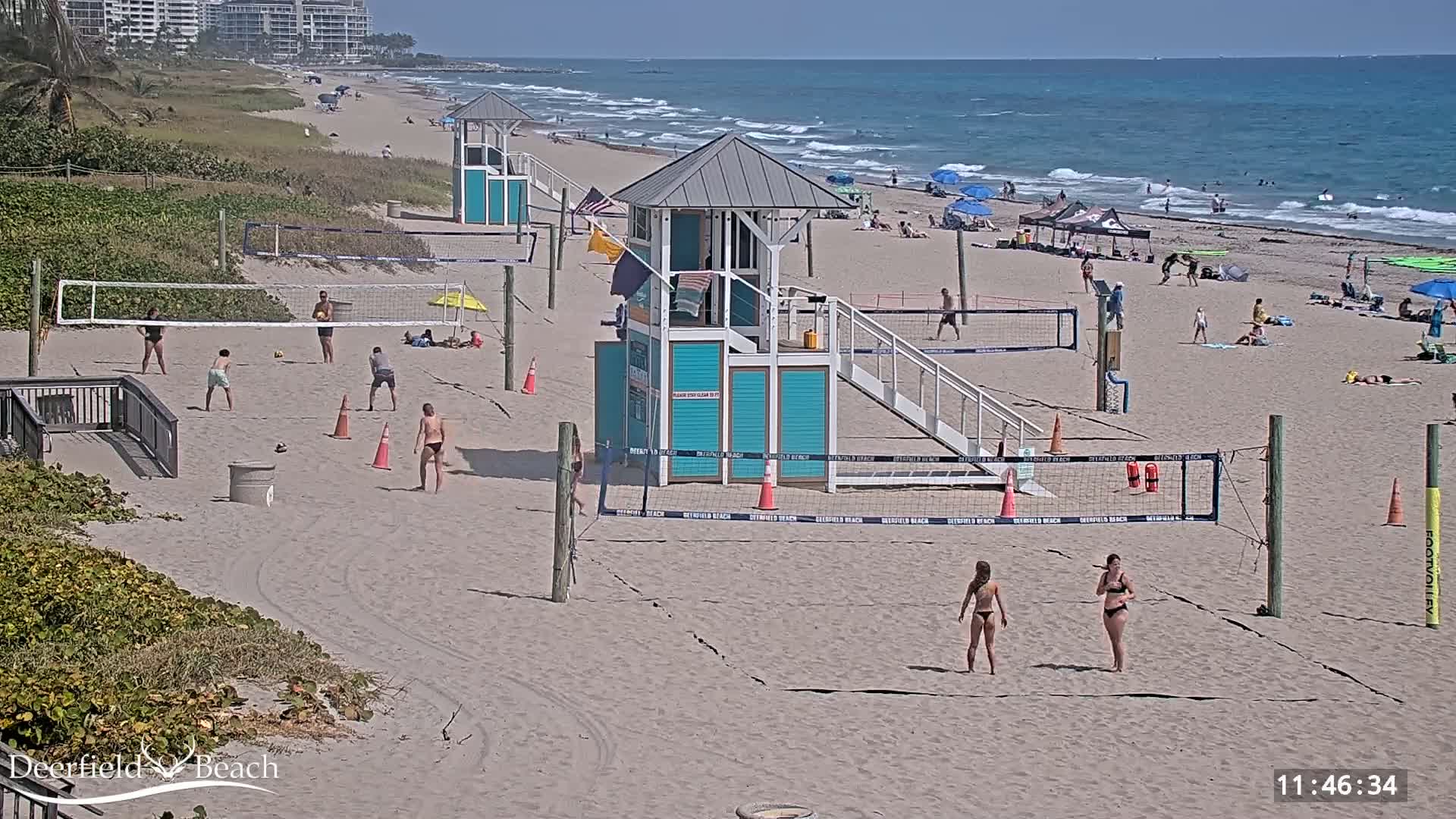 A sunny beach scene shows people playing volleyball on several courts near lifeguard stands, with a calm ocean in the background.
