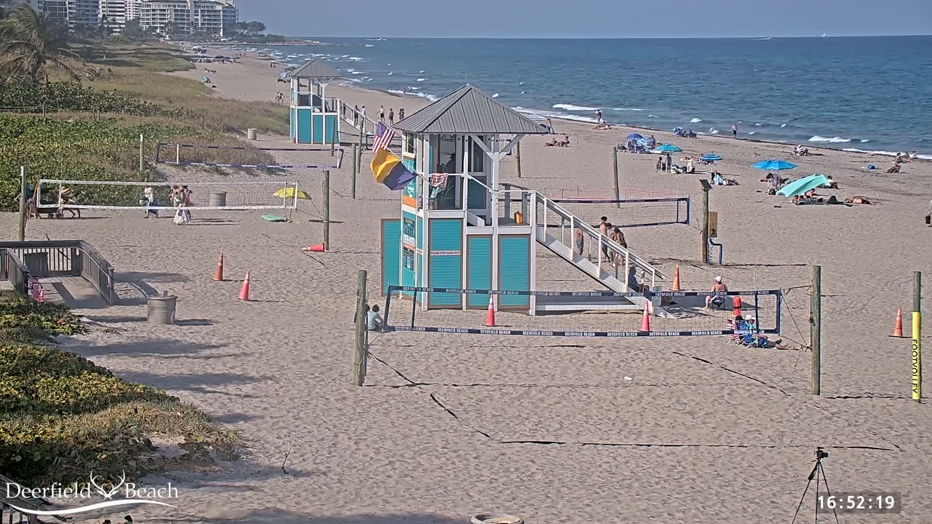 A sandy beach with lifeguard stands, volleyball nets, and scattered beachgoers under a clear, sunny sky, overlooks a calm ocean.