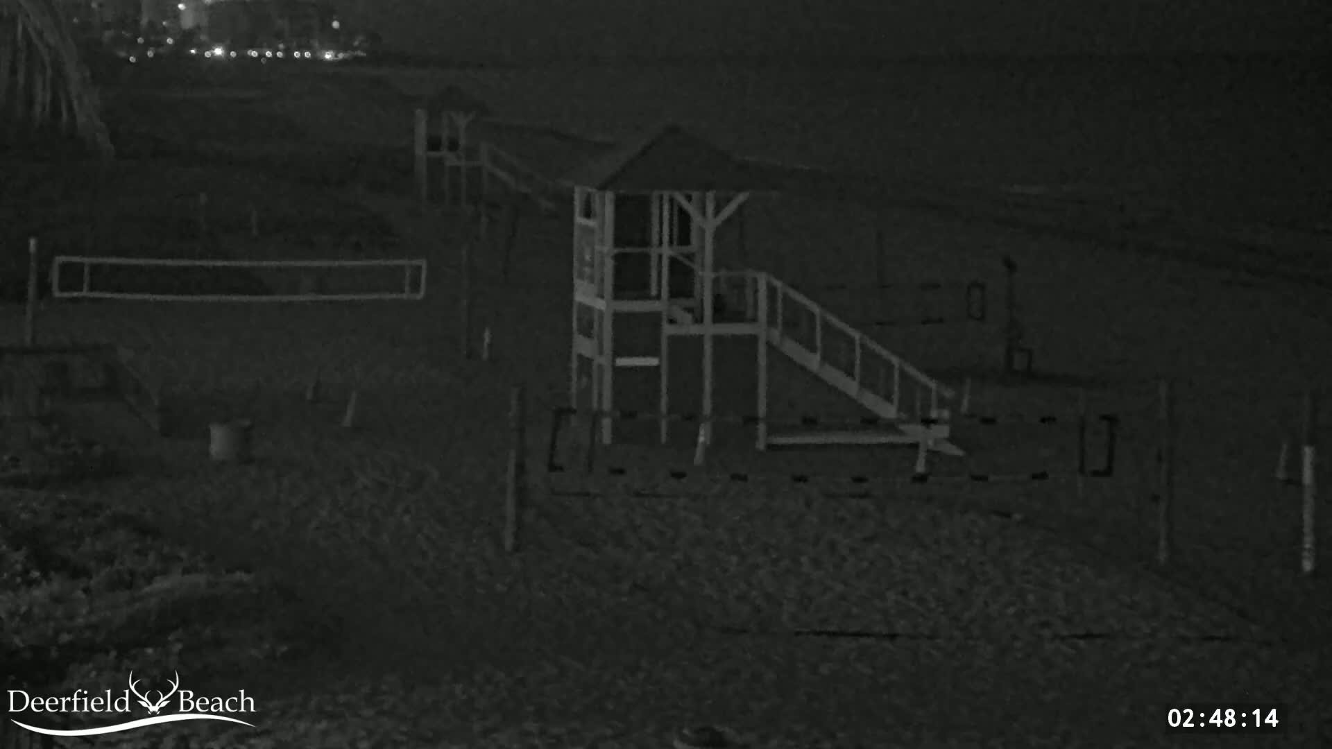 A nighttime, low-light image shows a beach volleyball net and a small elevated platform with a ramp and stairs on a sandy beach.