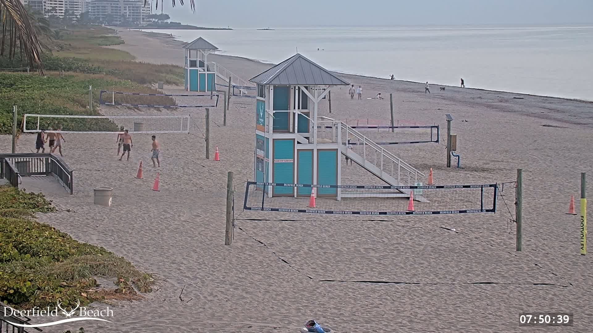 A sandy beach with several volleyball courts, lifeguard stands, and a few people playing volleyball and walking along the shoreline under overcast skies.