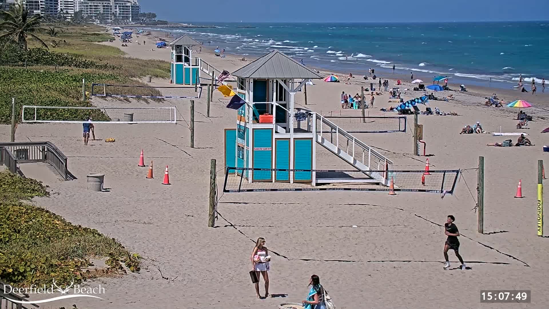 A sandy beach with volleyball courts, lifeguard stands, and several people enjoying a sunny day with moderately wavy ocean waters.